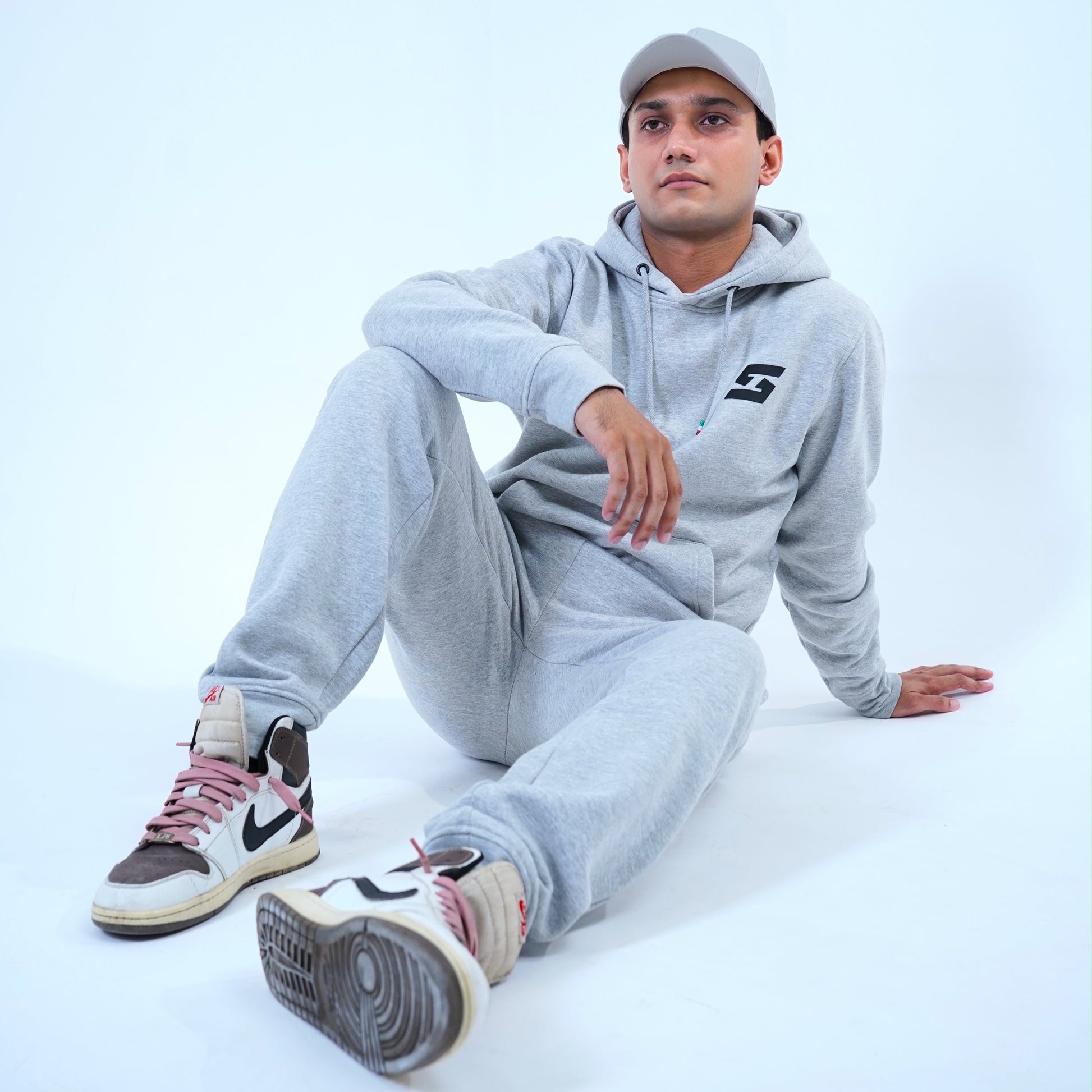 Man wearing a gray jogger tracksuit with pullover hoodie and sweatpants, styled with sneakers and a cap, sitting casually.