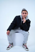 Man wearing a winter hooded jacket and casual street fashion, sitting on a stool in a stylish pose against a white background.